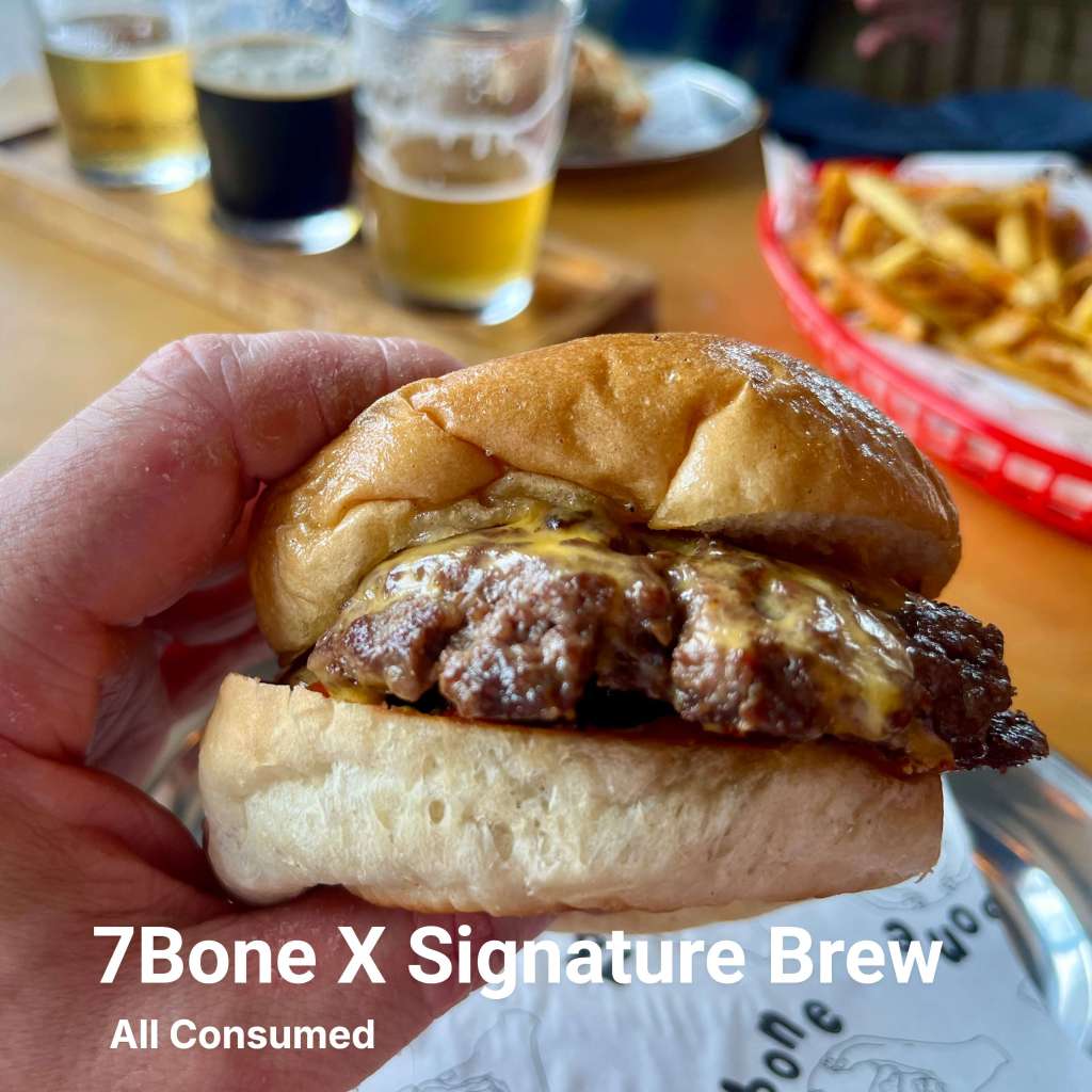 A close-up of a hand holding a 7Bone Burger with melted cheese on a soft bun, with glasses of beer in the background.